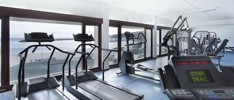 A gym with treadmills, ellipticals, and weight machines, featuring large windows with a view of the beach.