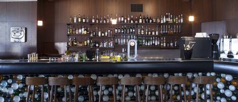 A bar area with a wooden bar counter, multiple shelves of liquor bottles, a coffee machine, and a wall of colorful glass bottles.