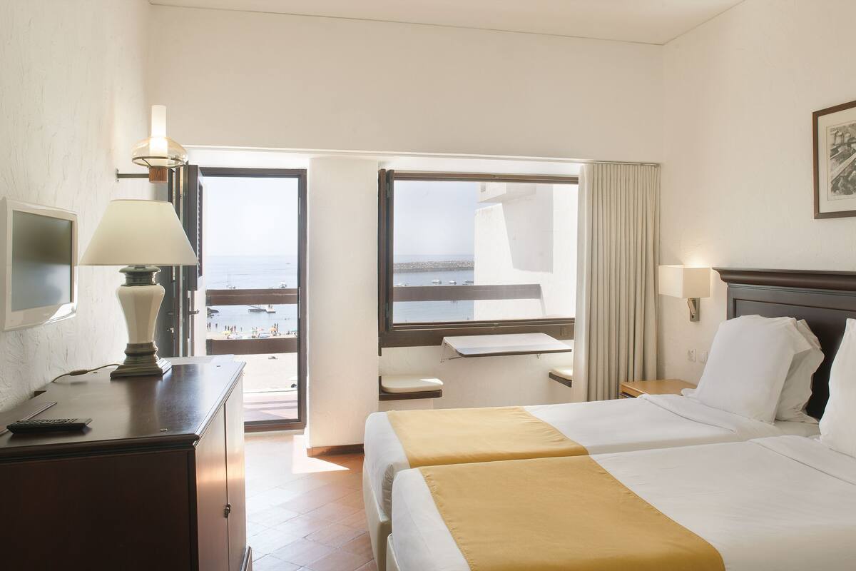 A hotel room with two beds, a desk, a lamp, a TV, and a view of the beach.