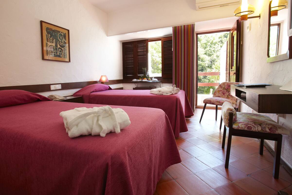 A hotel room with two beds, a desk, and a chair. There is a window with shutters, a painting on the wall, and a lamp on the desk.