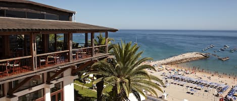 A beachside hotel with a balcony overlooking the sea, a palm tree, and beach umbrellas.