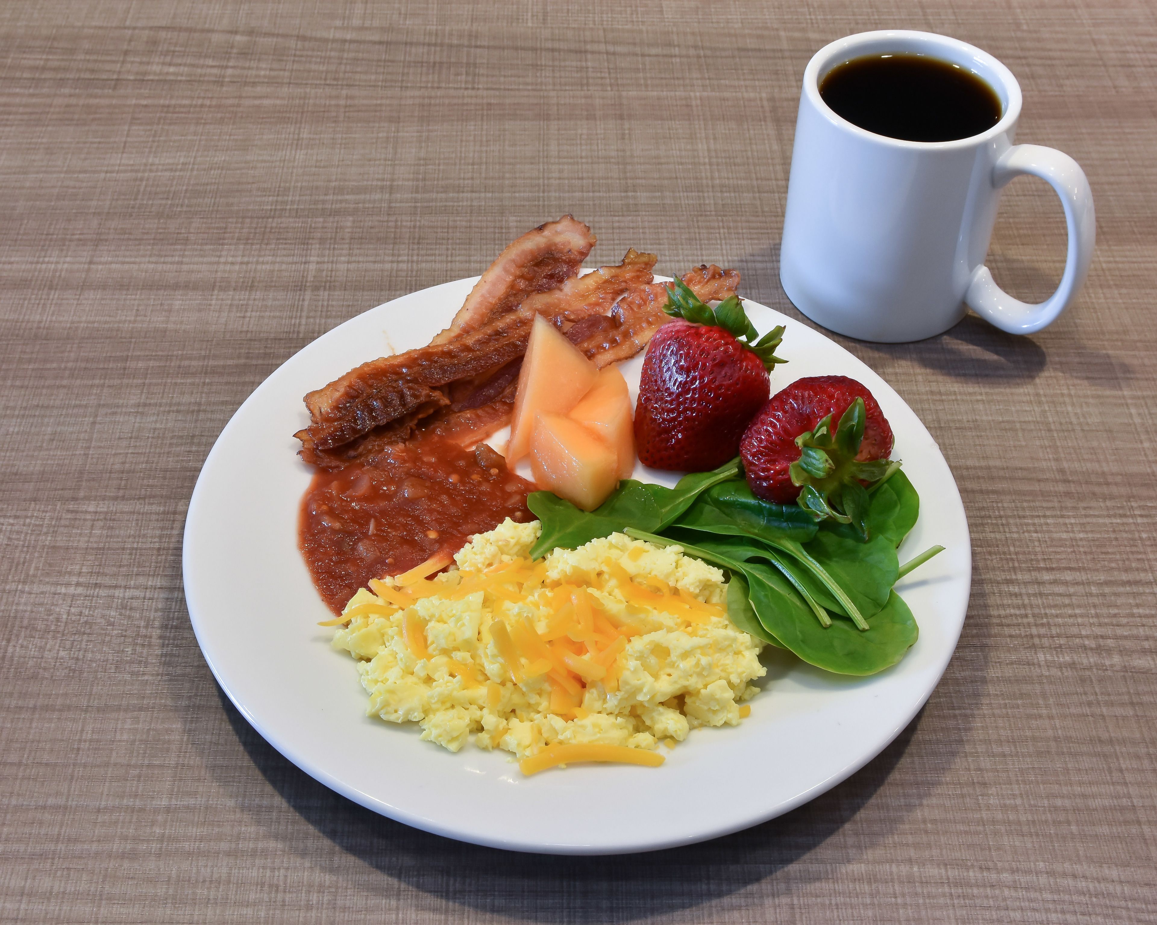 Un plato con huevos revueltos, tocino, fresas, espinacas y una taza de café sobre una mesa.