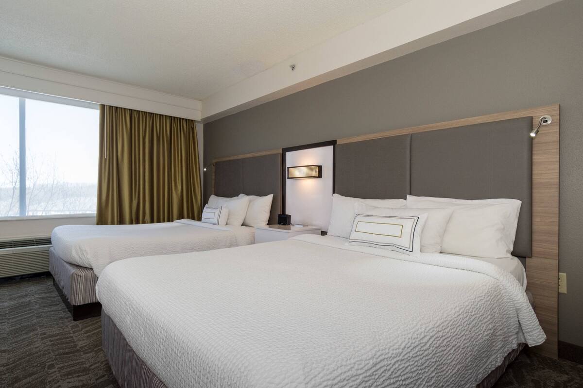 Habitación de hotel con dos camas, una mesita de noche, una lámpara y una ventana con cortinas.