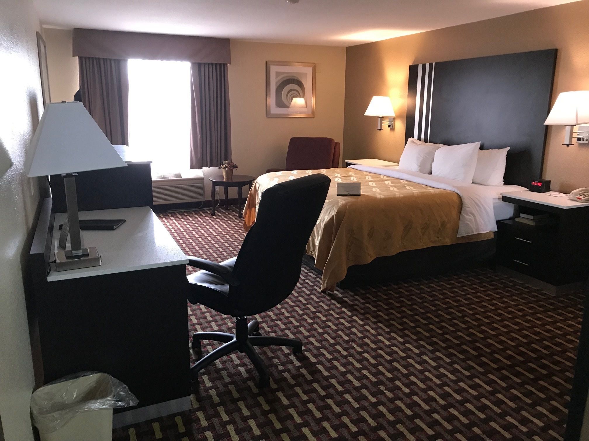 A hotel room with a bed, desk, chair, lamps, and a clock.