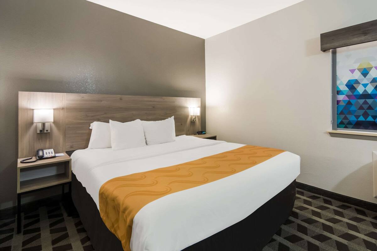 A hotel room with a large bed, two bedside tables with lamps, a wooden headboard, and a window with a geometric pattern.