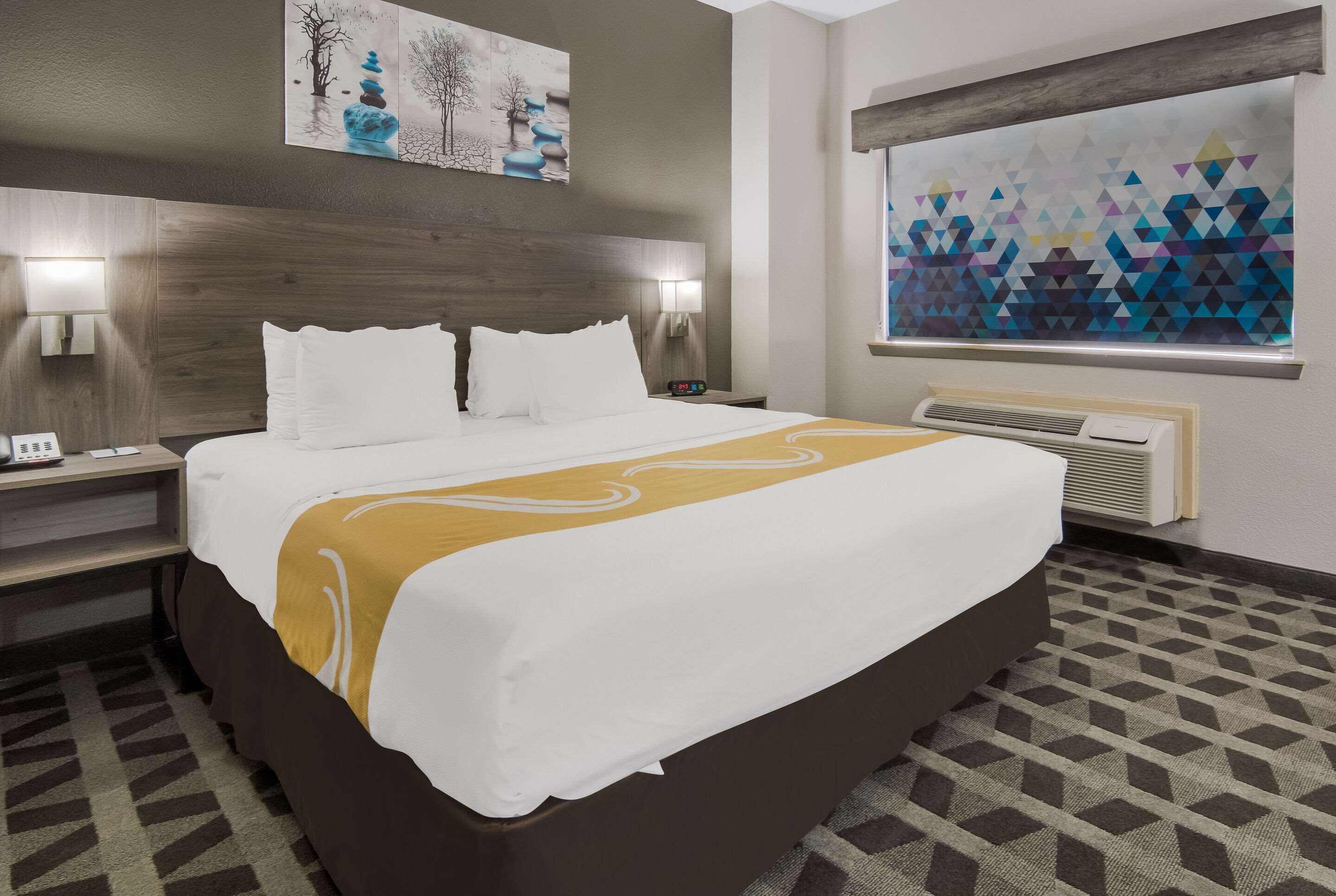 Una habitación de hotel con una cama grande, dos mesitas de noche con lámparas, un aire acondicionado de pared y una obra de arte sobre la cama.