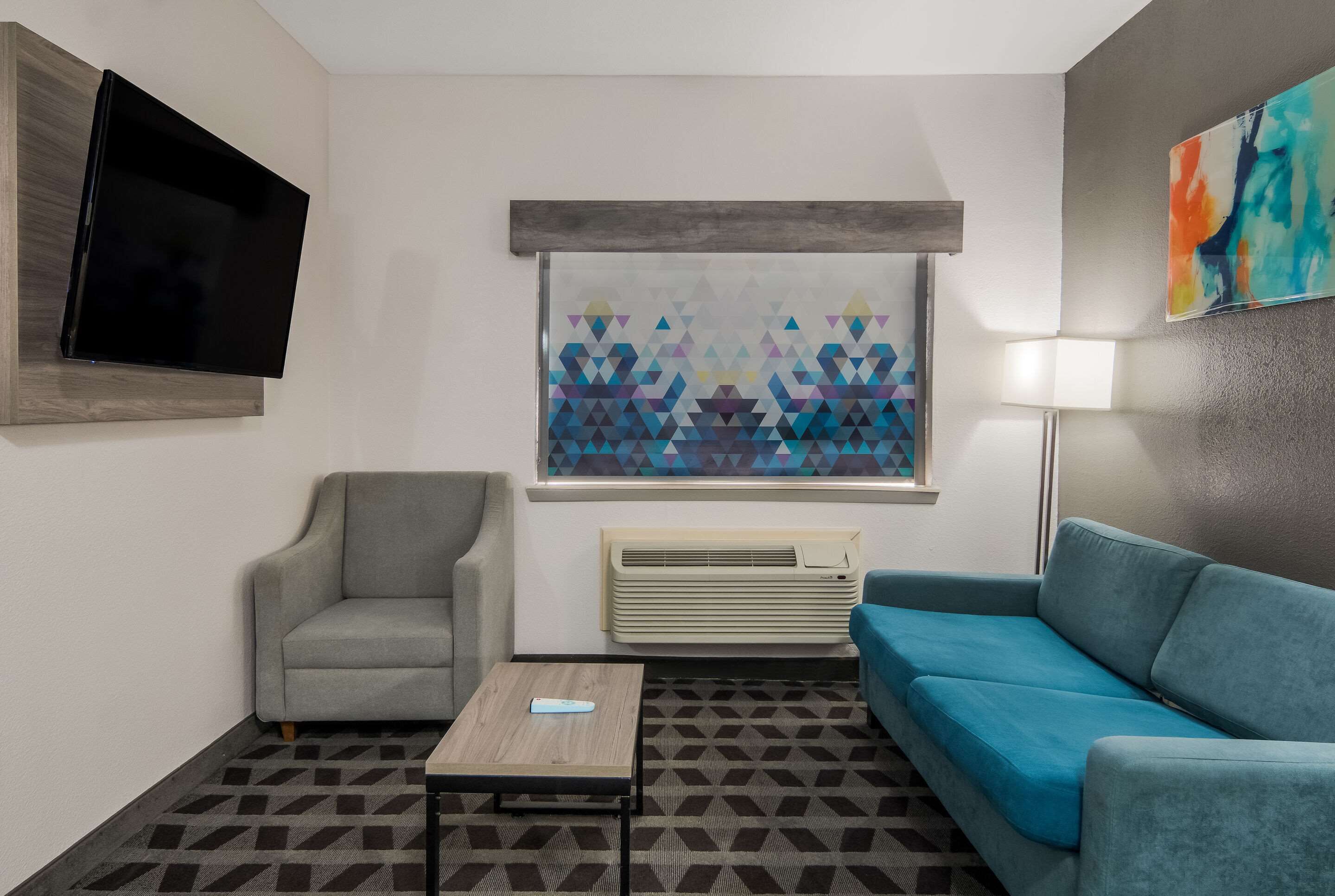 A modern living room with a grey armchair, a blue sofa, a coffee table, a flat-screen TV mounted on the wall, a window with a geometric patterned blind, and a wall-mounted air conditioner.