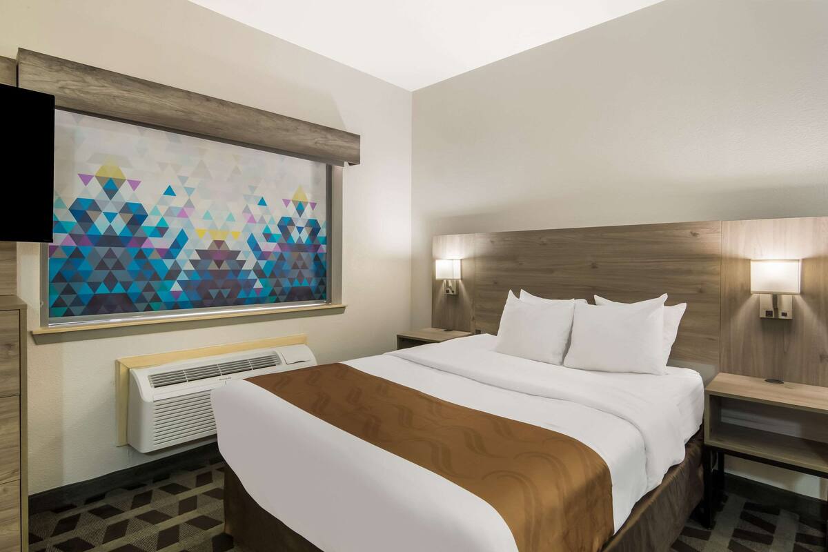 A hotel room with a large bed, a wall-mounted TV, a geometric patterned artwork, a wooden headboard, bedside lamps, and a built-in air conditioning unit.