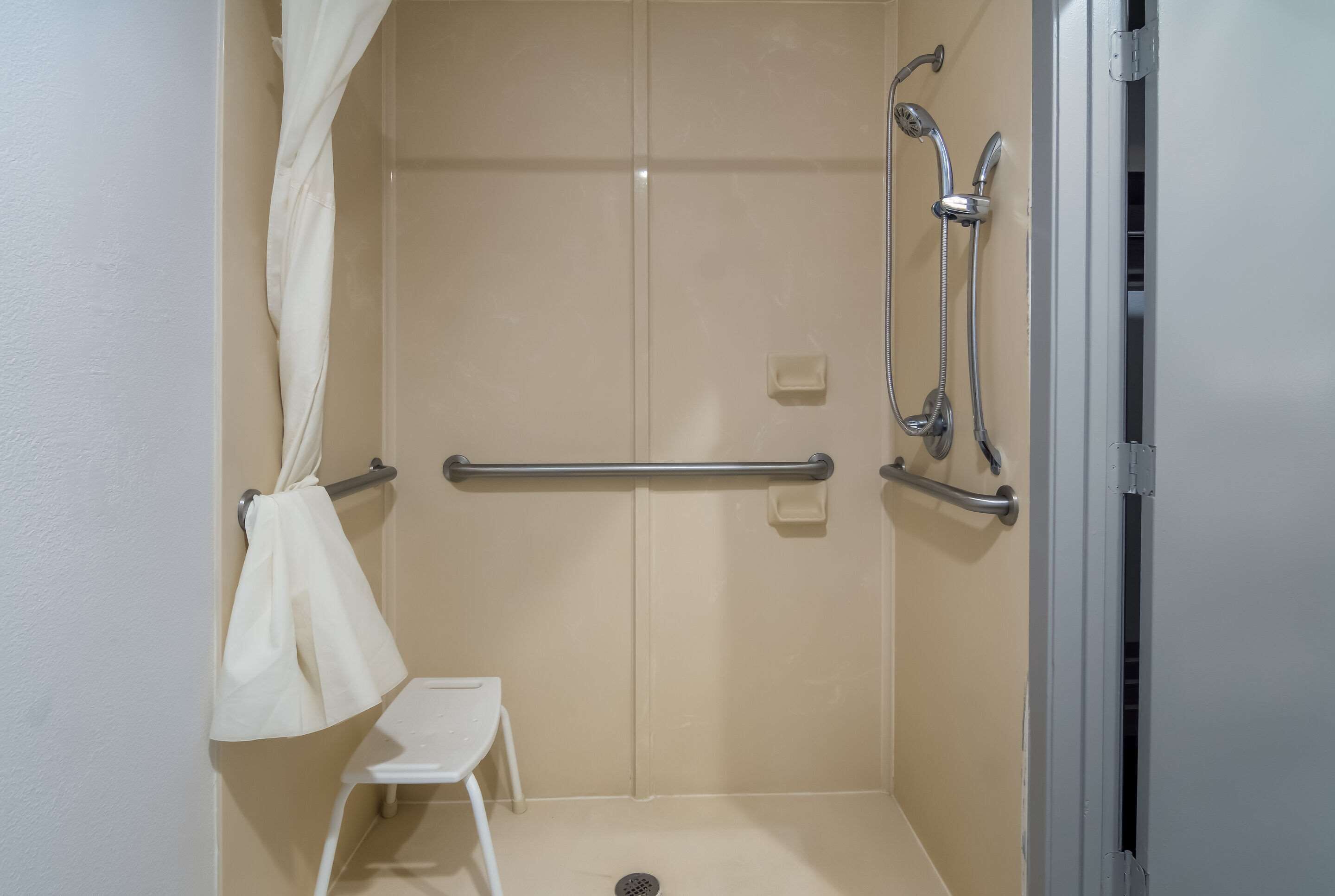 A small, enclosed shower area with a white shower chair, grab bars, and a showerhead.
