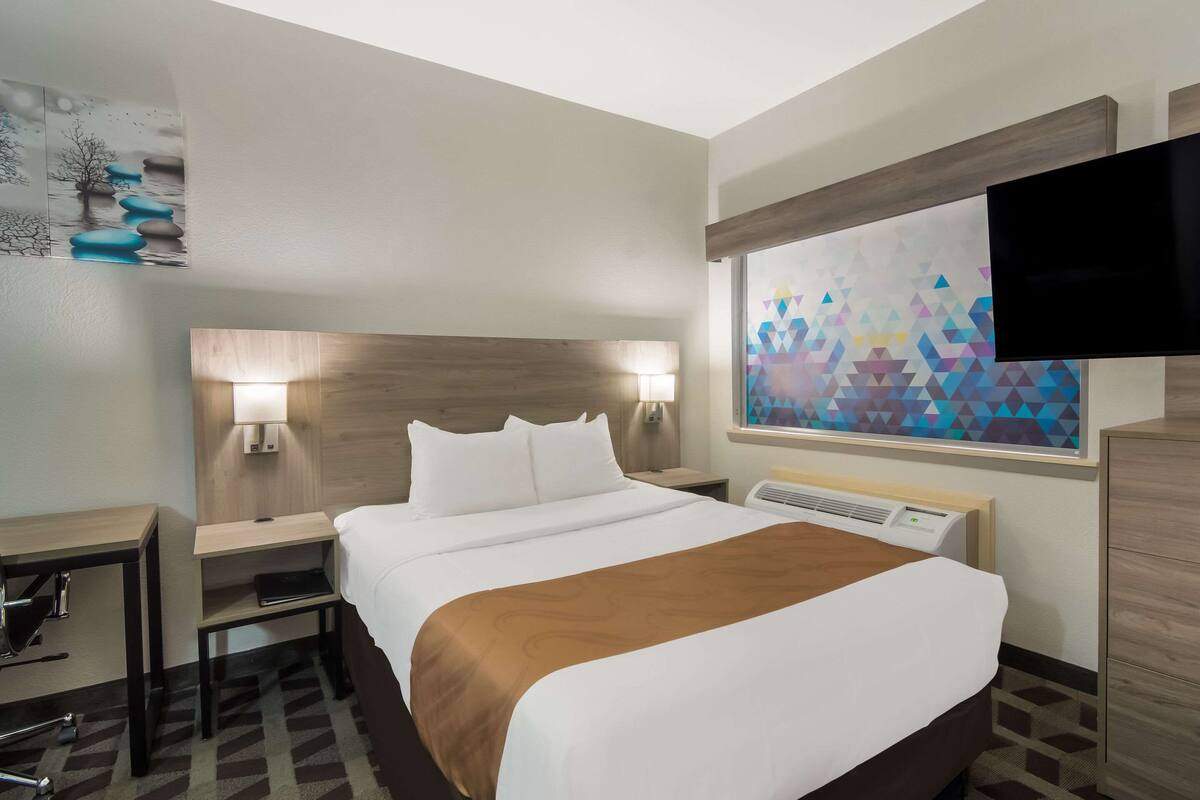 A hotel room with a bed, bedside tables, a desk, a chair, a TV, and a painting on the wall.
