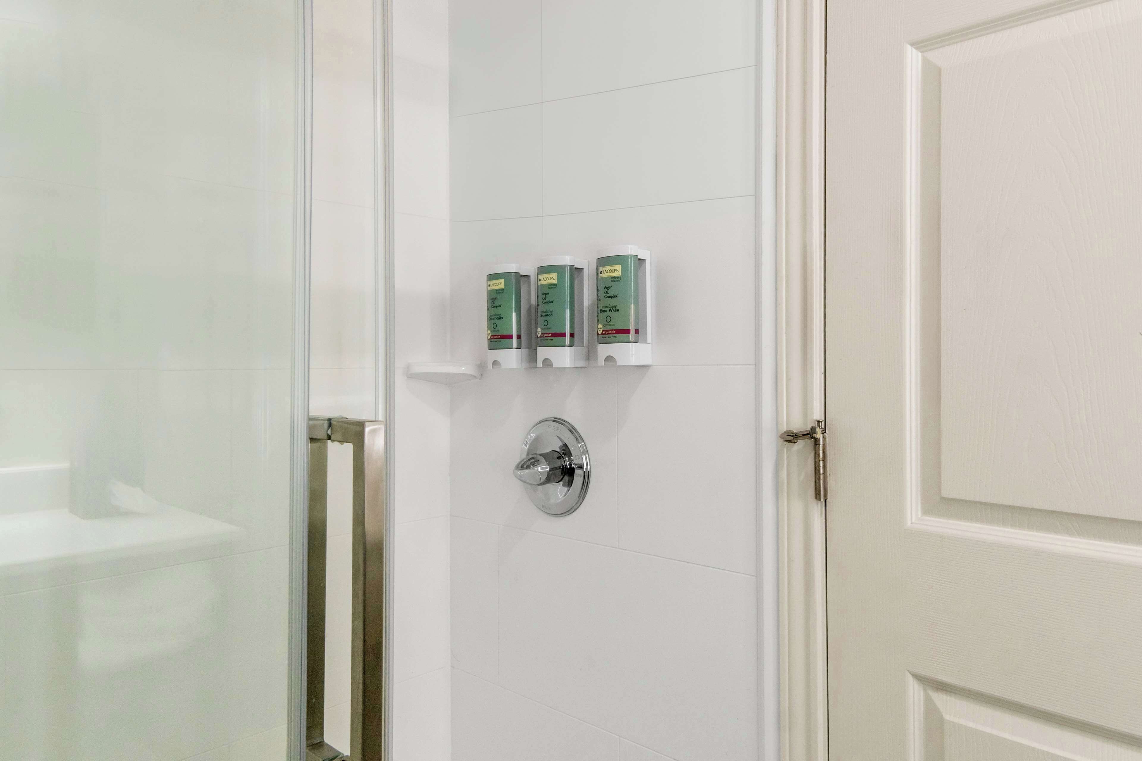 Um banheiro com chuveiro de vidro, parede com azulejos brancos e três dosadores verdes em uma prateleira.