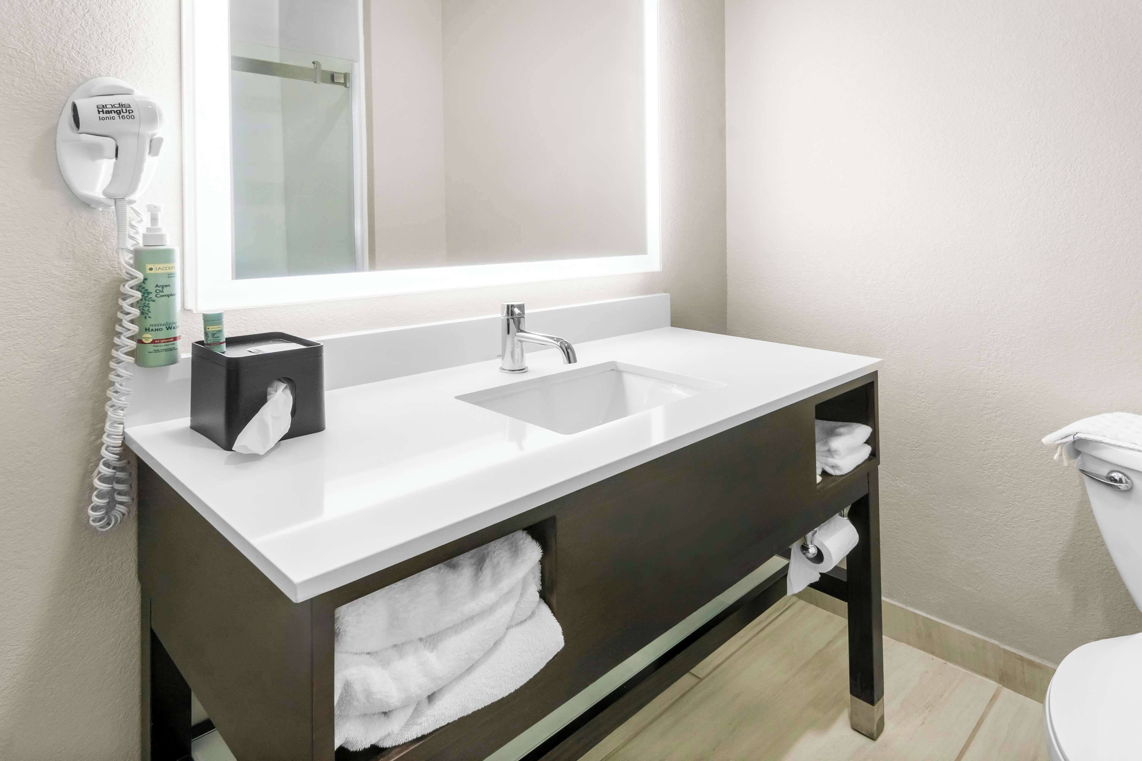 A modern bathroom with a white sink, a mirror with integrated lighting, a hairdryer, and a toilet.