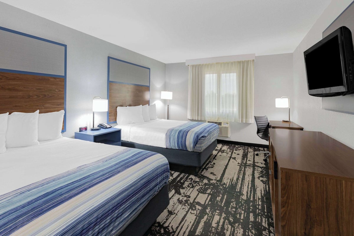 A hotel room with two beds, a desk, a chair, a flat-screen TV, and a large window with curtains.