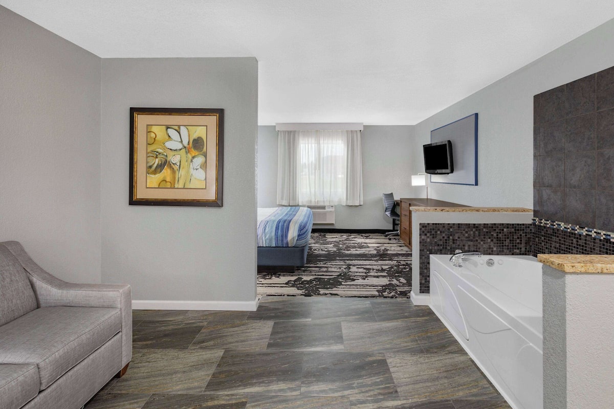 A modern hotel room with a grey sofa, a bed with a striped blanket, a large window with sheer curtains, a flat-screen TV mounted on the wall, and a bathtub with a mosaic tile surround.