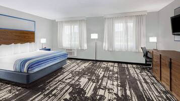 Habitación de hotel con cama, escritorio, silla, dos ventanas con cortinas, televisor de pantalla plana y alfombra estampada.