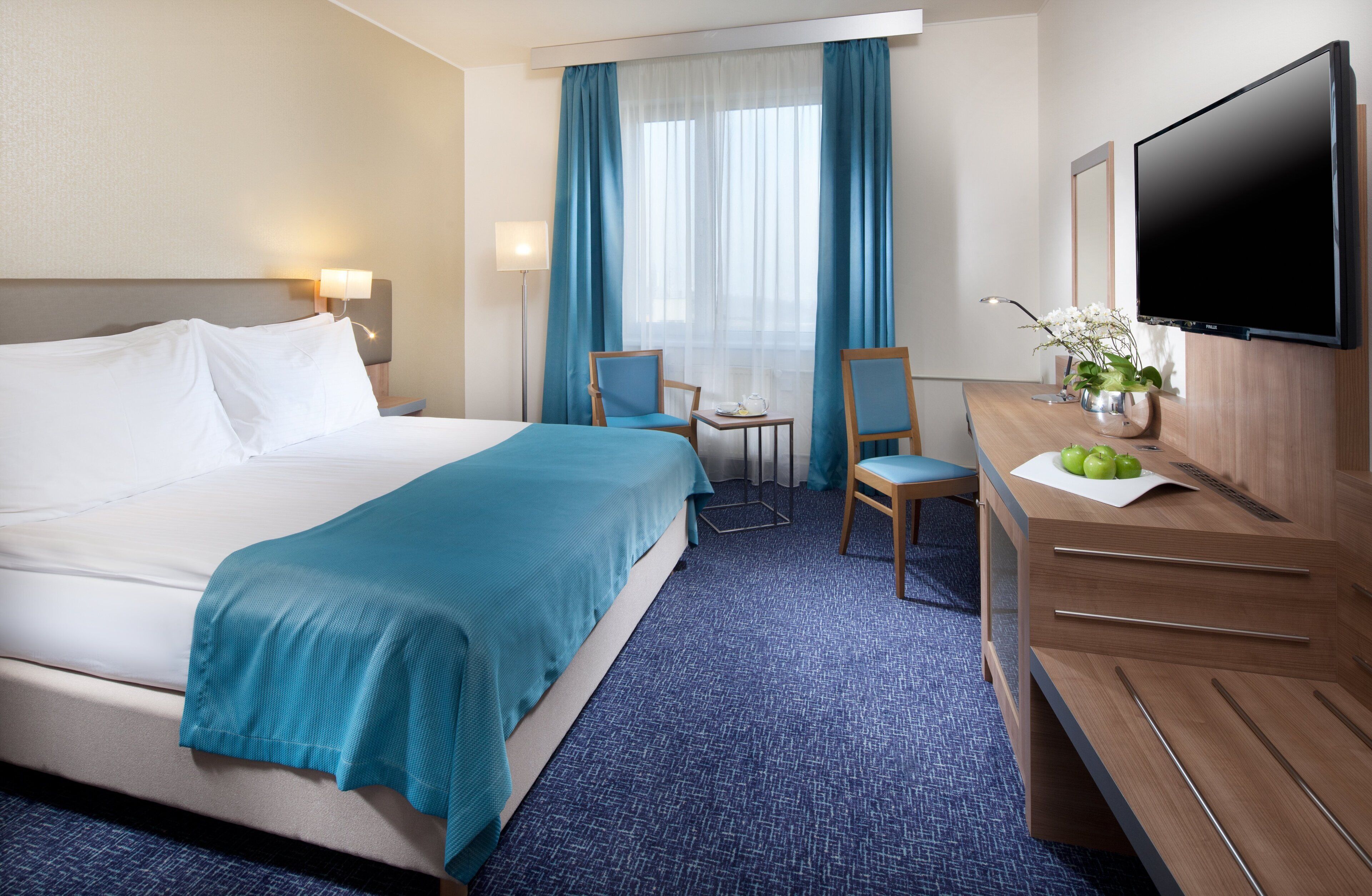 Habitación de hotel con una cama grande, un escritorio con televisión de pantalla plana, una silla, una mesita con un florero y una ventana con cortinas.