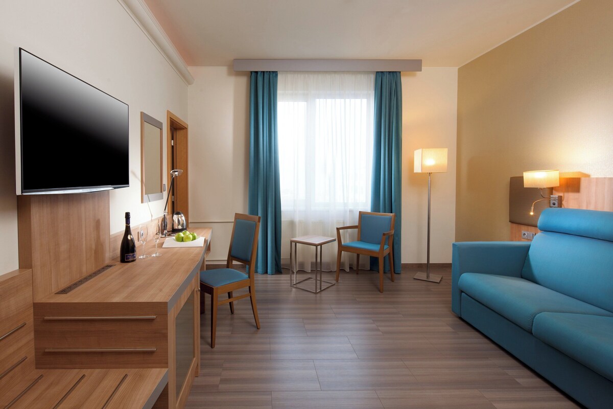 A hotel room with a flat-screen TV, a wooden desk with a bottle and glasses, two chairs, a sofa, and a floor lamp.