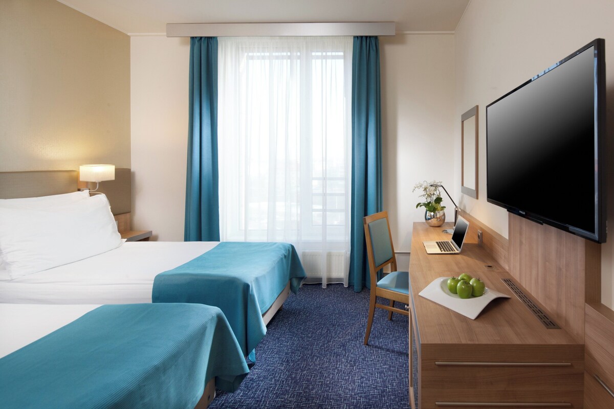 Een hotelkamer met twee bedden, een bureau met een laptop, een flatscreen televisie en een raam met gordijnen.