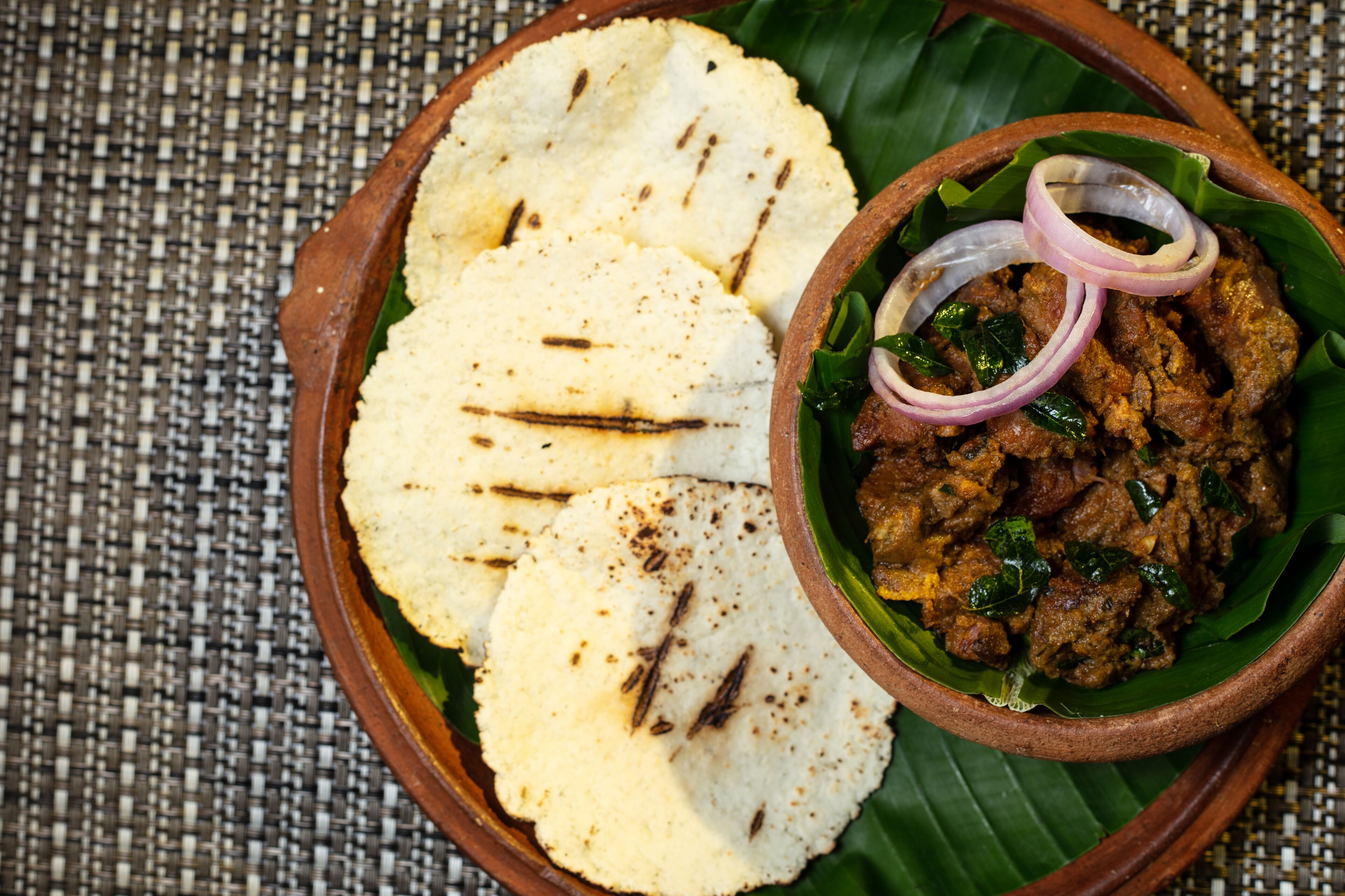 A plate with flatbreads and a bowl of meat dish, served on a banana leaf.