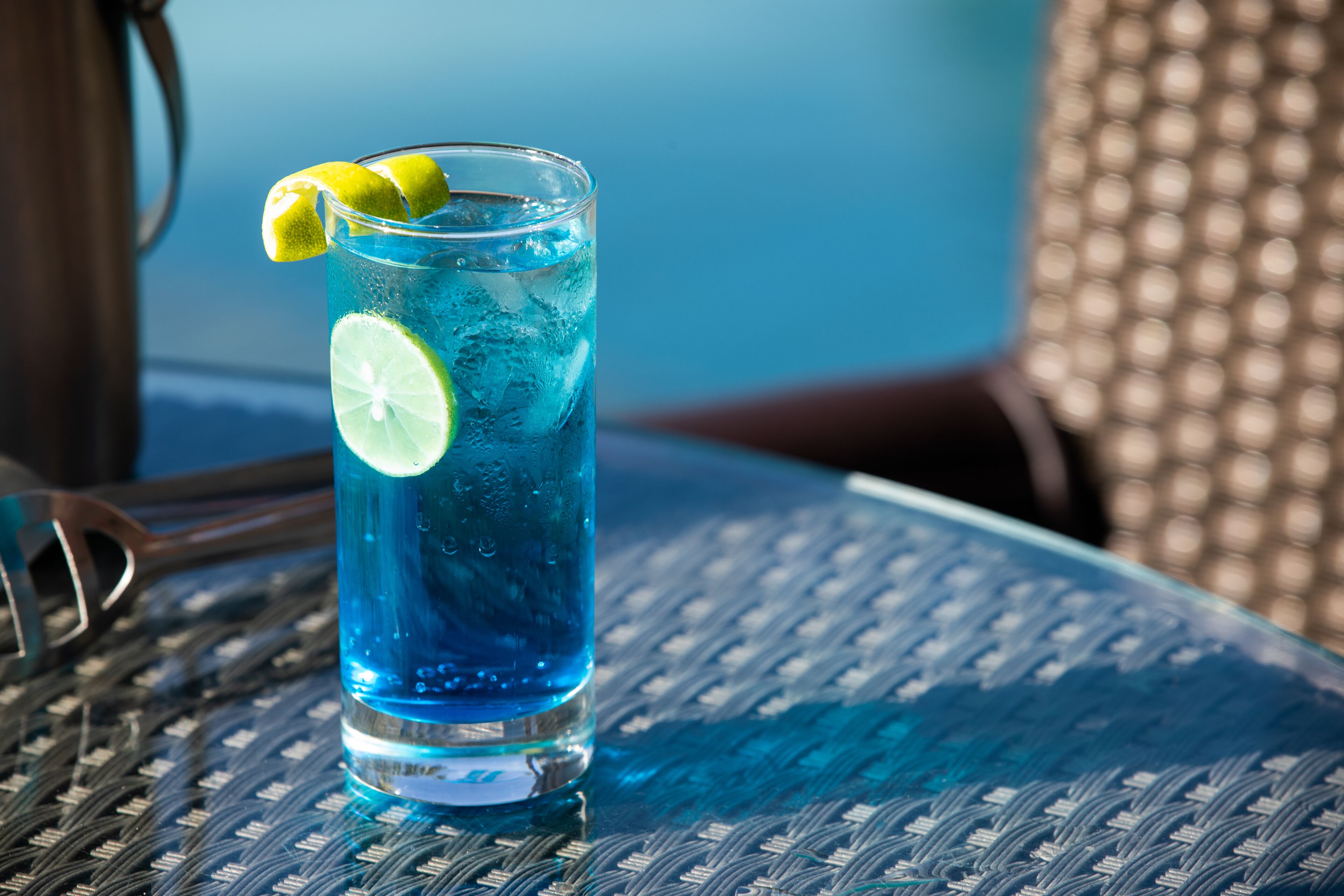A tall glass with blue liquid and lime slices, placed on a woven table.