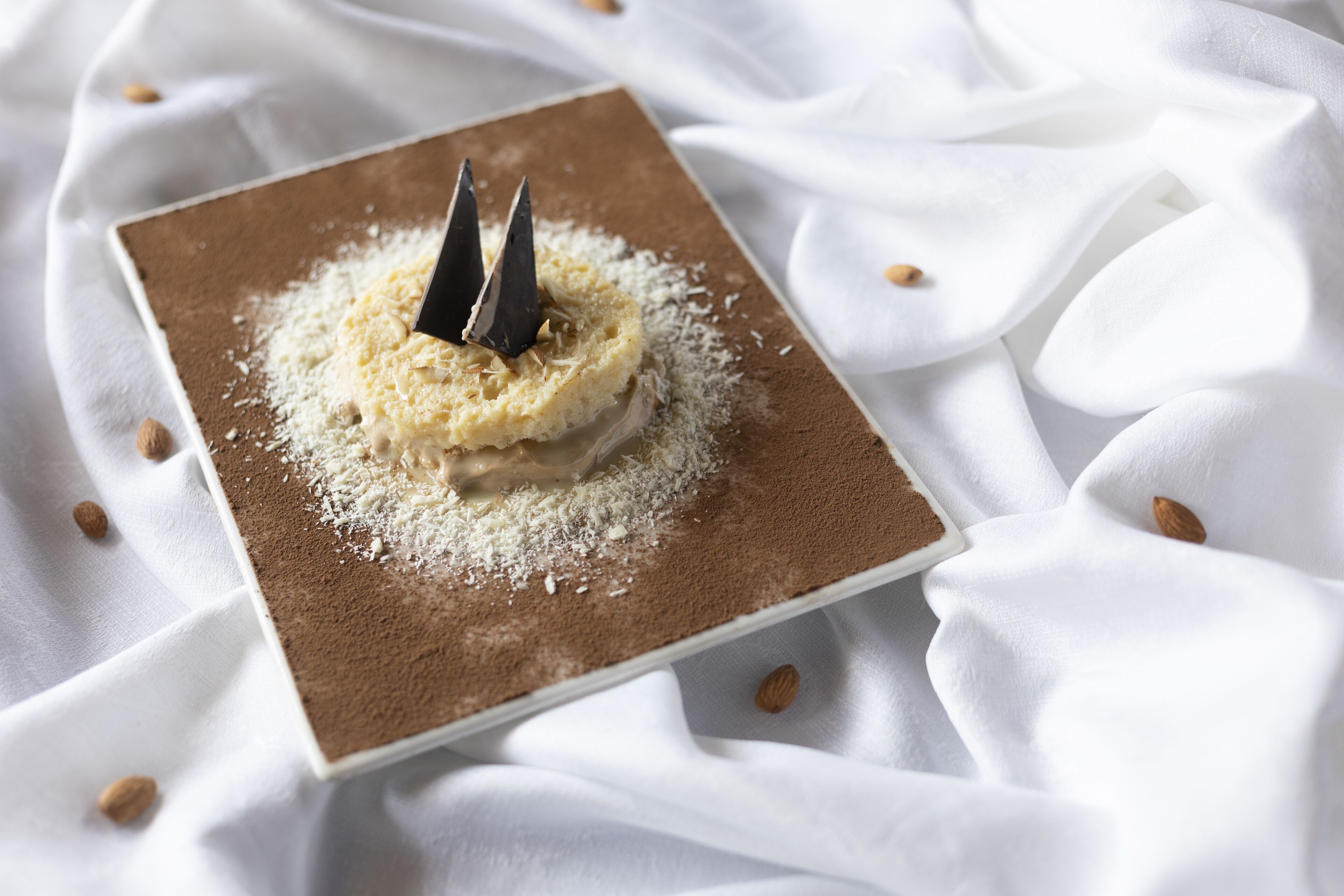 Un postre en un plato cuadrado con base marrón, rodeado de tela blanca y almendras.