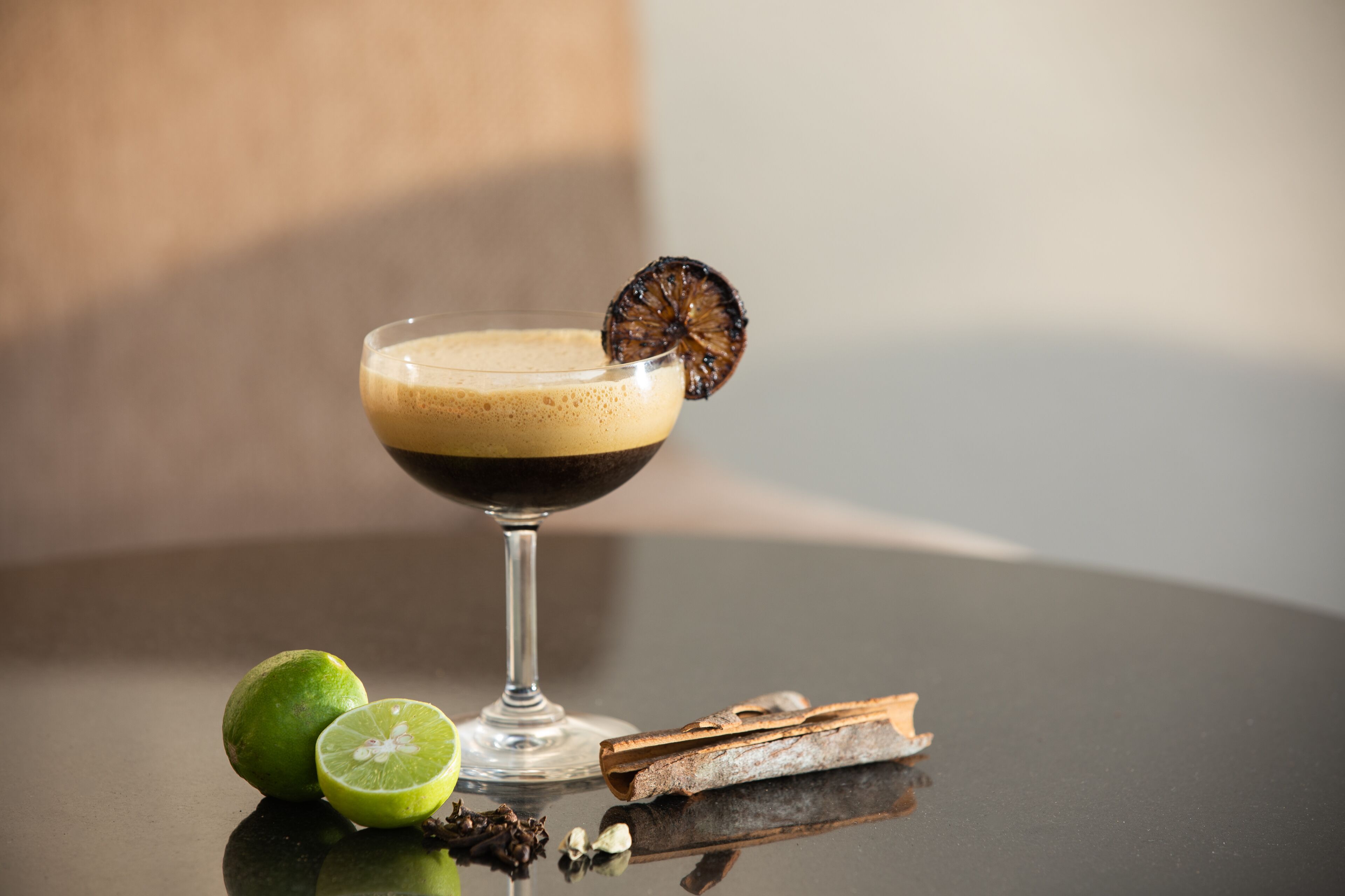 Una copa de cóctel con un líquido oscuro y una rodaja de fruta deshidratada en el borde, acompañada de media lima y palitos de canela sobre una superficie reflectante.