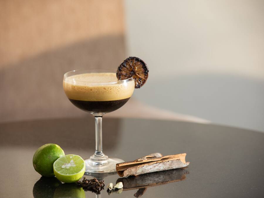 Una copa de cóctel con un líquido oscuro y una rodaja de fruta deshidratada en el borde, acompañada de media lima y palitos de canela sobre una superficie reflectante.