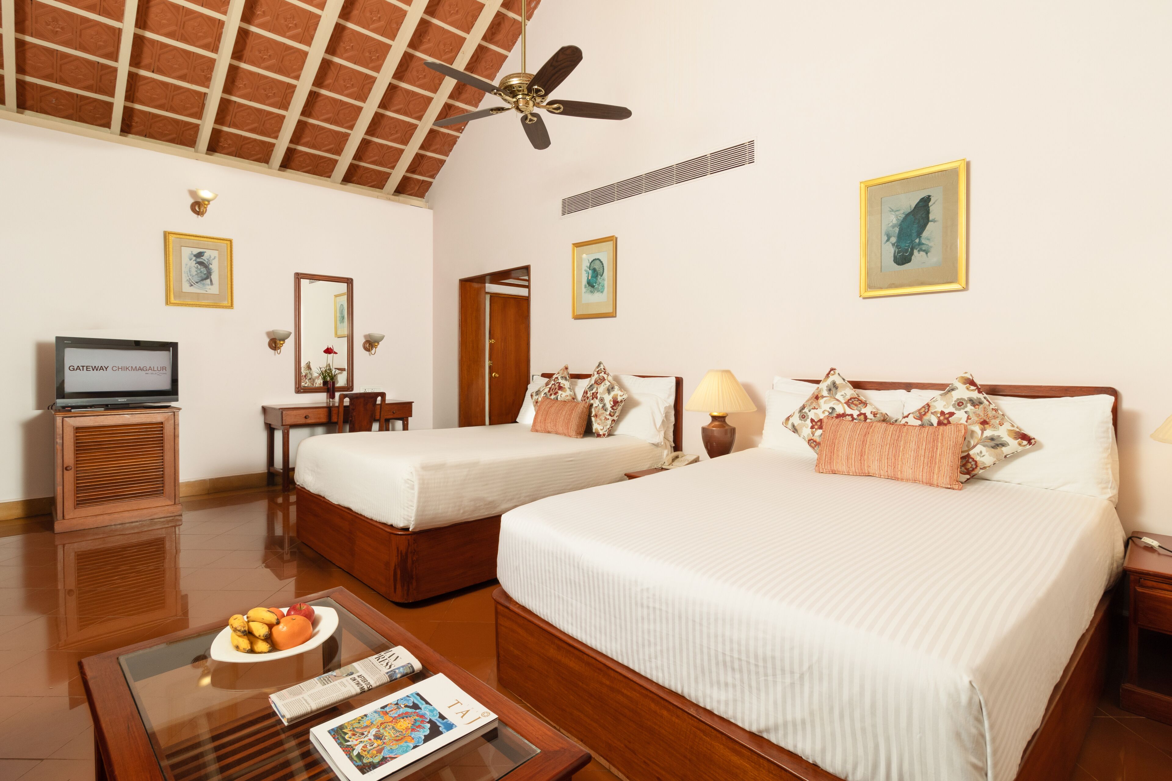 Habitación de hotel con dos camas individuales, ventilador de techo, televisor, mesa de comedor con fruta y obras de arte en las paredes.
