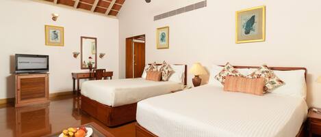 Habitación de hotel con dos camas individuales, ventilador de techo, televisor, mesa de comedor con fruta y obras de arte en las paredes.