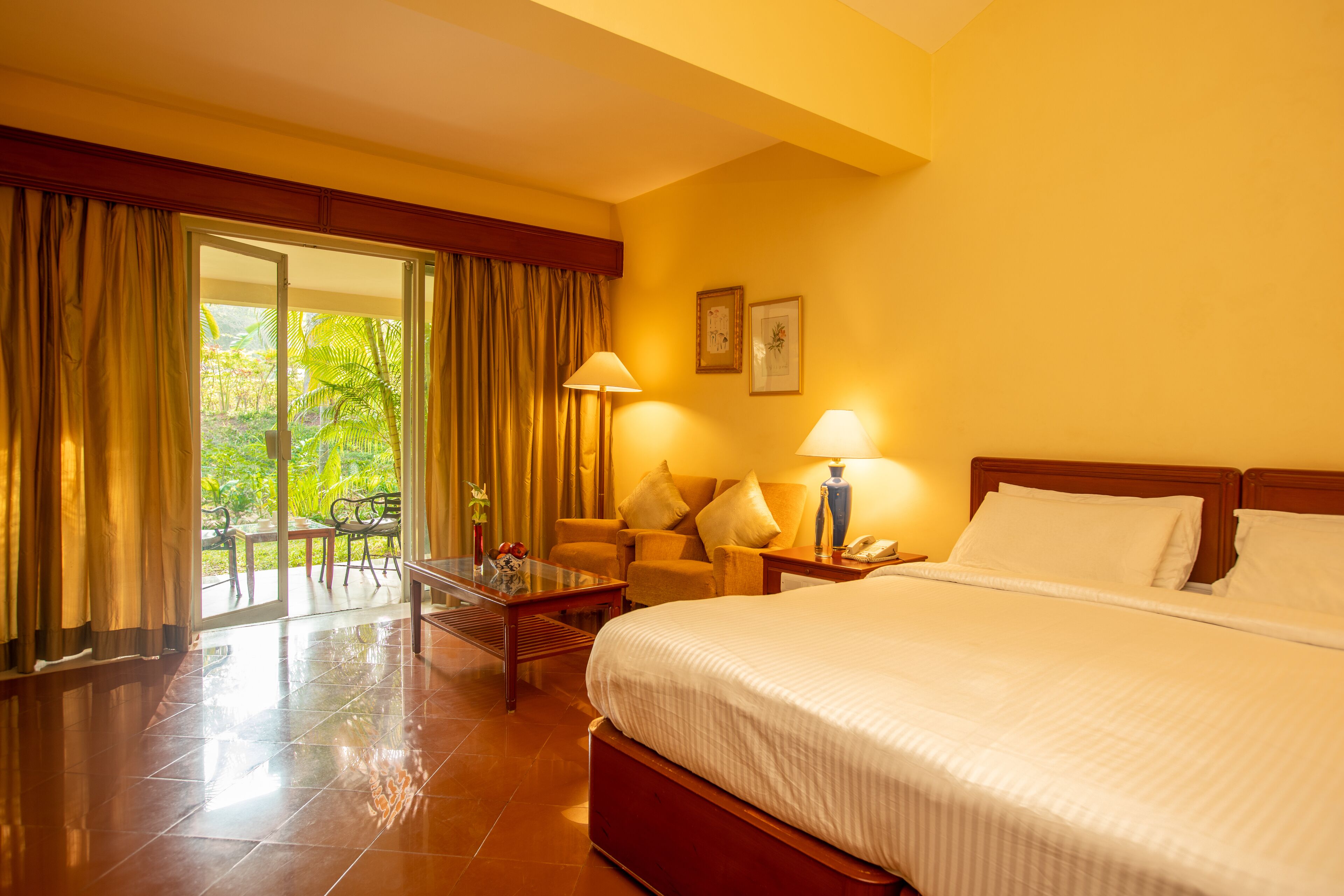 A hotel room with a large bed, two bedside lamps, a nightstand, a glass table with a vase, and a view of greenery through a sliding door.