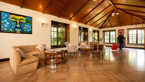 A spacious living room with a high wooden ceiling, large windows, and a beige sofa. - Gateway Chikmagalur (Chikkamagaluru)