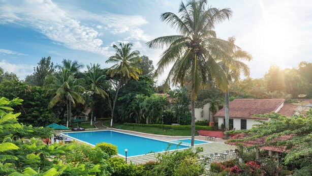 A swimming pool surrounded by lush greenery and palm trees, with a view of a building with a red-tiled roof. - Gateway Chikmagalur (Chikkamagaluru)
