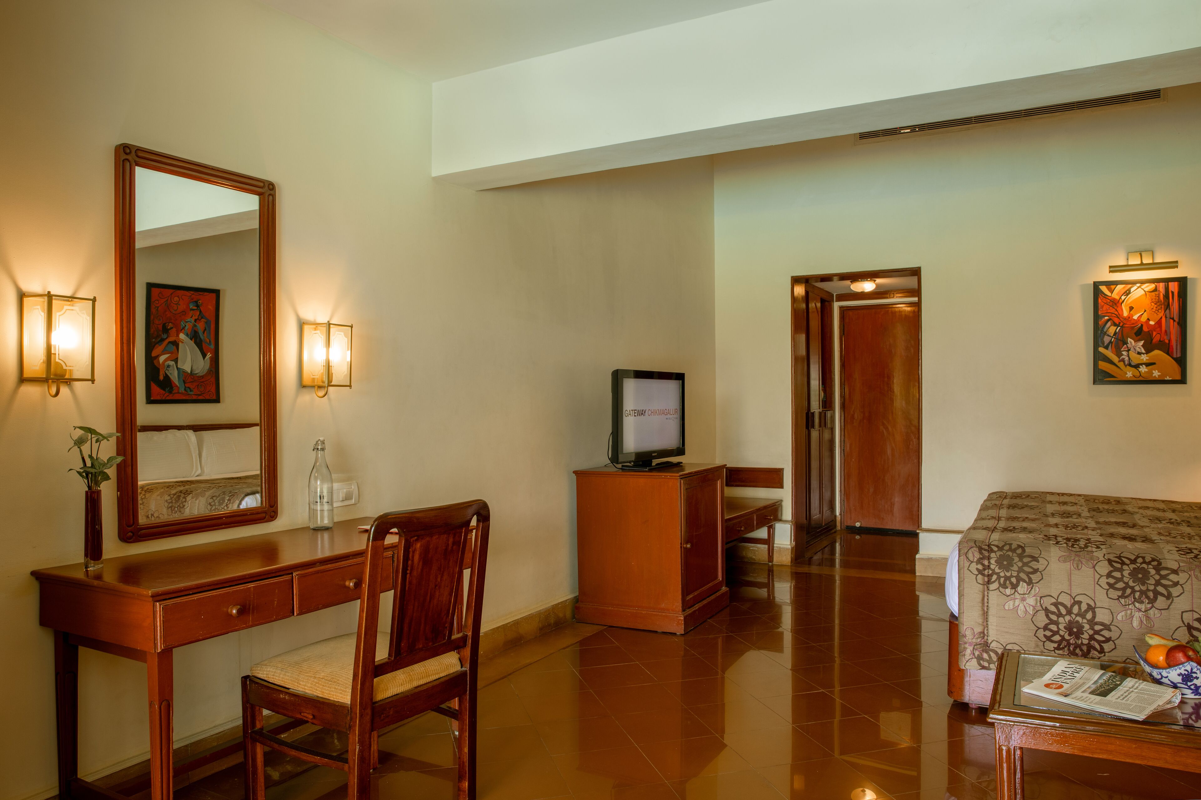 A hotel room with a wooden desk, a chair, a television, a bed with a floral bedspread, and a painting on the wall.