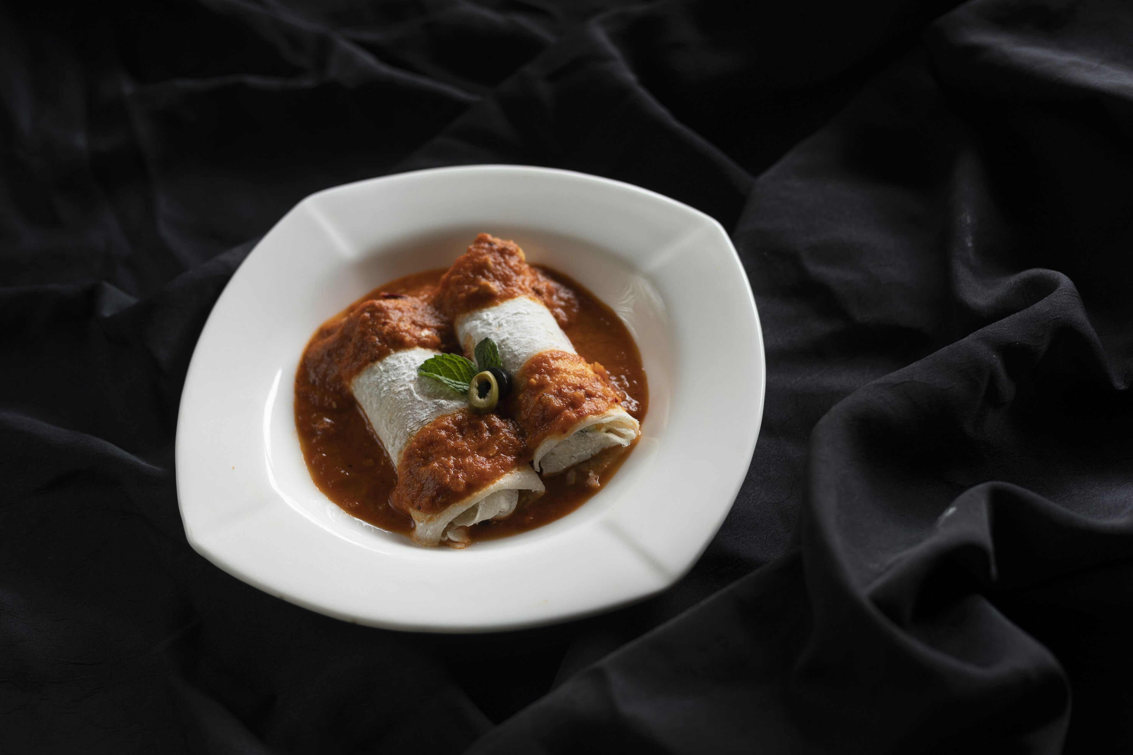 Un plato de enchiladas con salsa oscura, adornado con una hoja verde, sobre un plato blanco frente a un fondo de tela oscura.