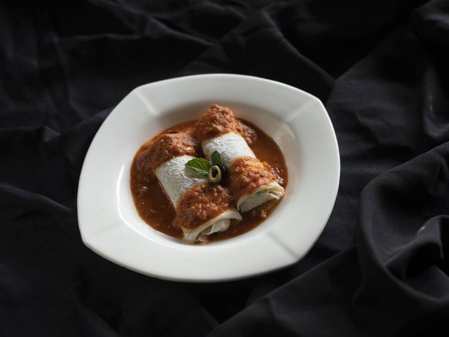 Un plato de enchiladas con salsa oscura, adornado con una hoja verde, sobre un plato blanco frente a un fondo de tela oscura.