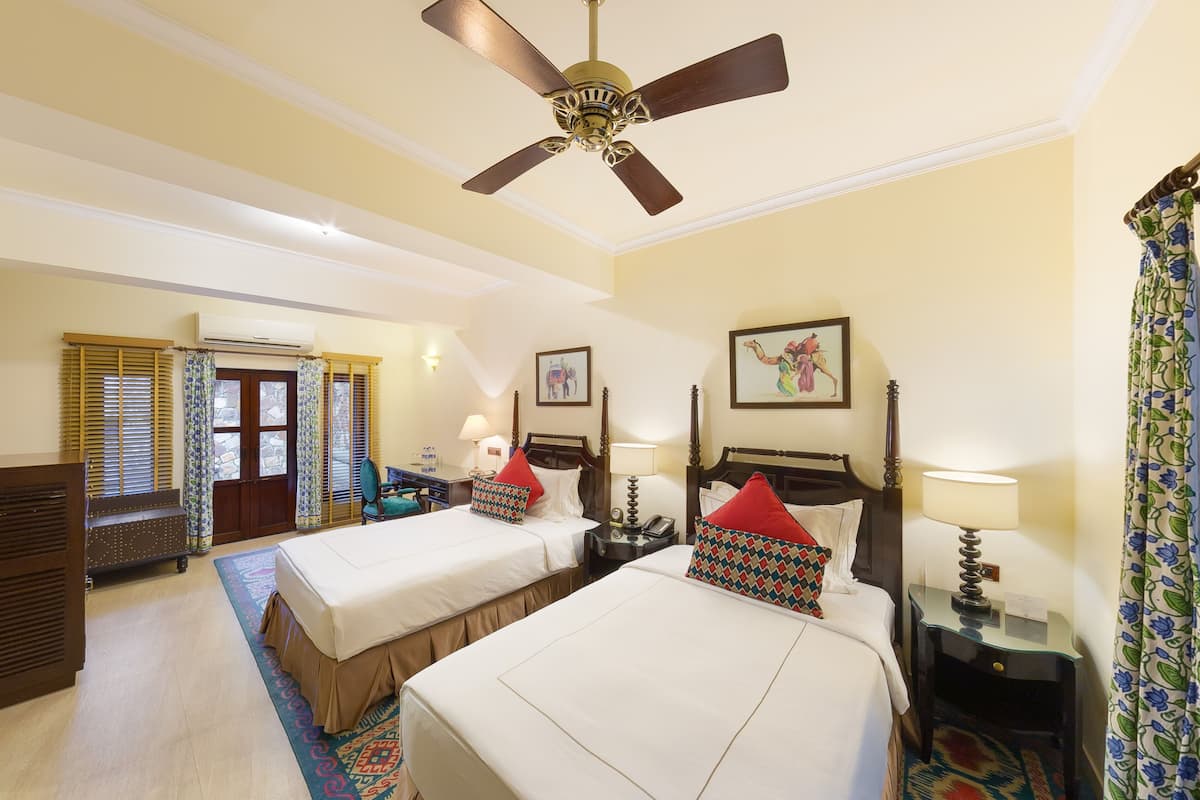 Une chambre d’hôtel avec deux lits, un ventilateur de plafond, un appareil de climatisation, une fenêtre avec des stores et un tapis à motifs.