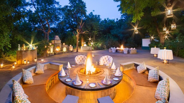 An outdoor dining area with a central fire feature, seating, and ambient lighting. - The Maharaja's Lodge, Ranthambore (Sawai Madhopur)