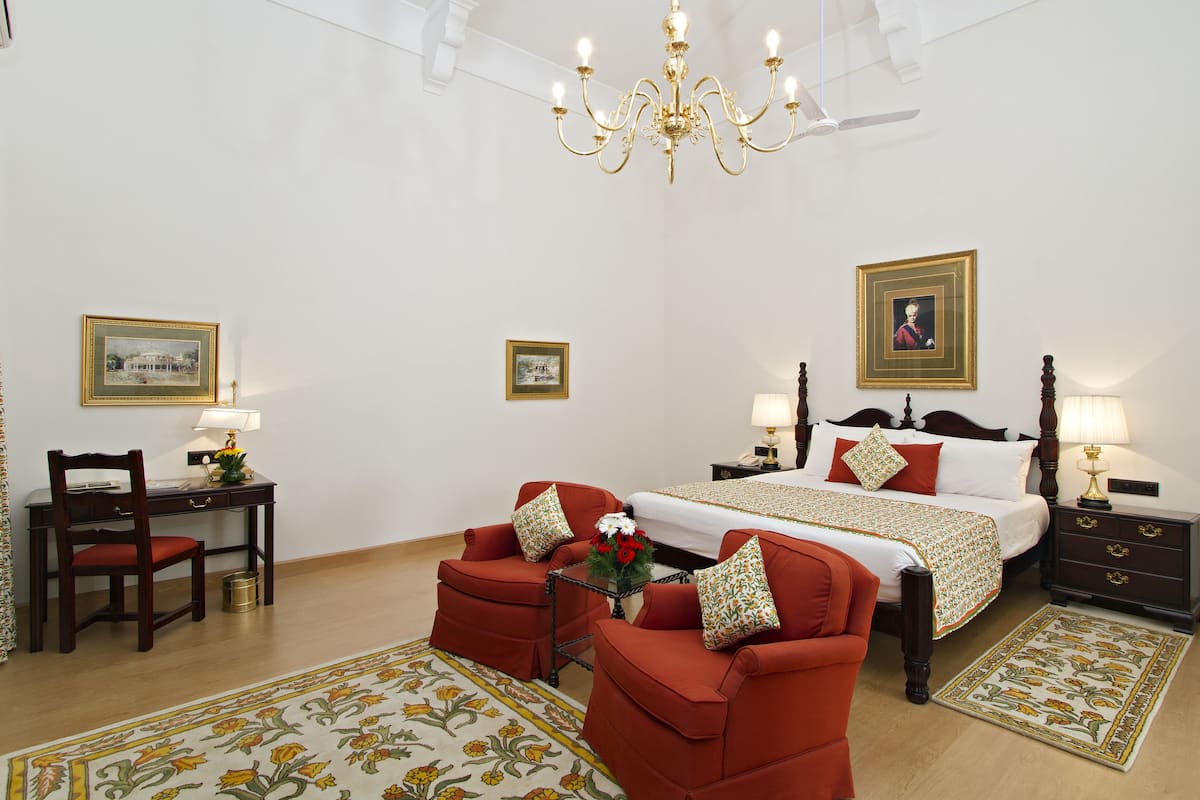 Une chambre d’hôtel avec un grand lit, deux fauteuils rouges, un bureau en bois et un lustre.