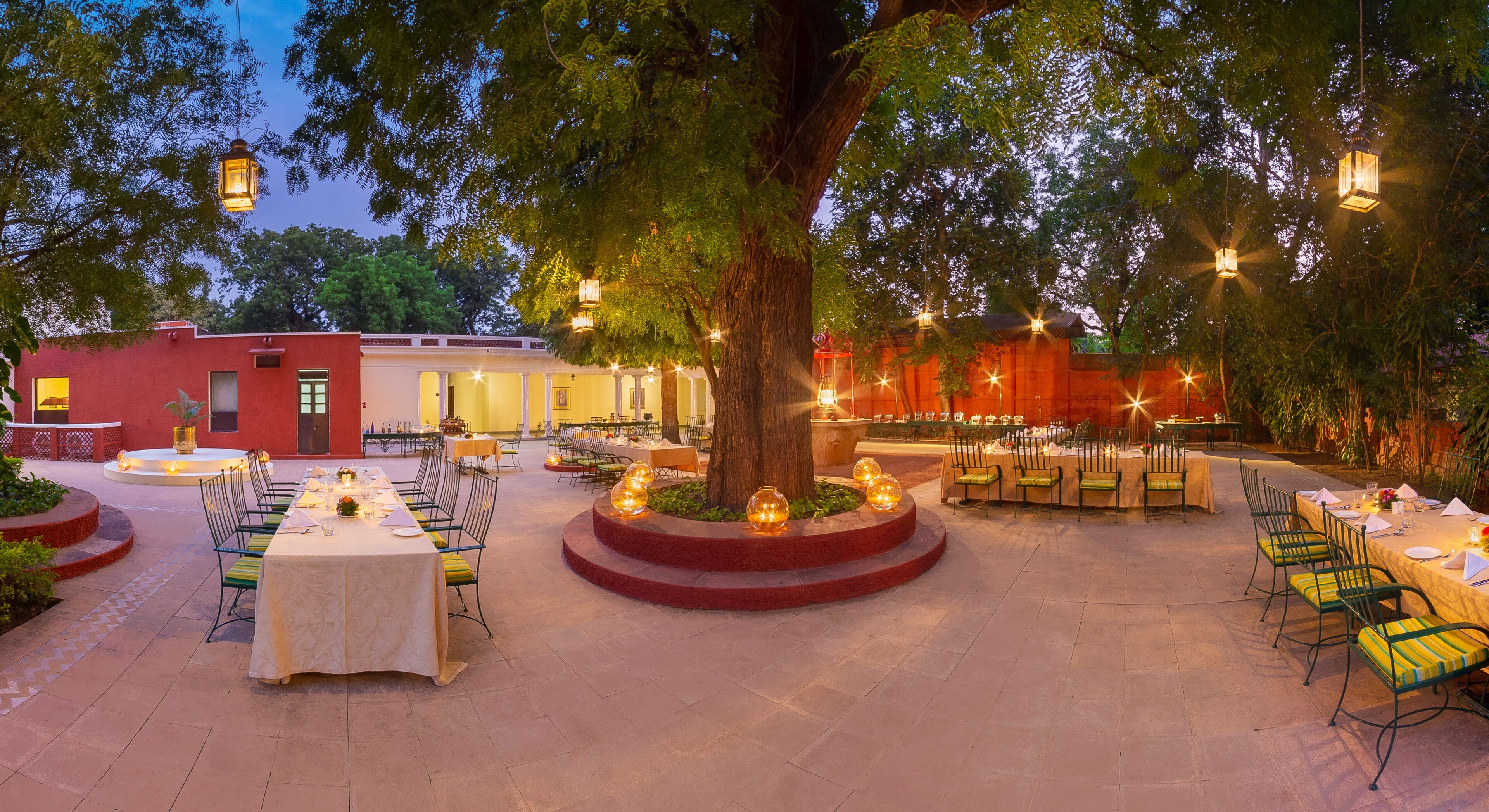 An outdoor dining area with round tables and chairs, surrounded by trees and illuminated by lanterns.