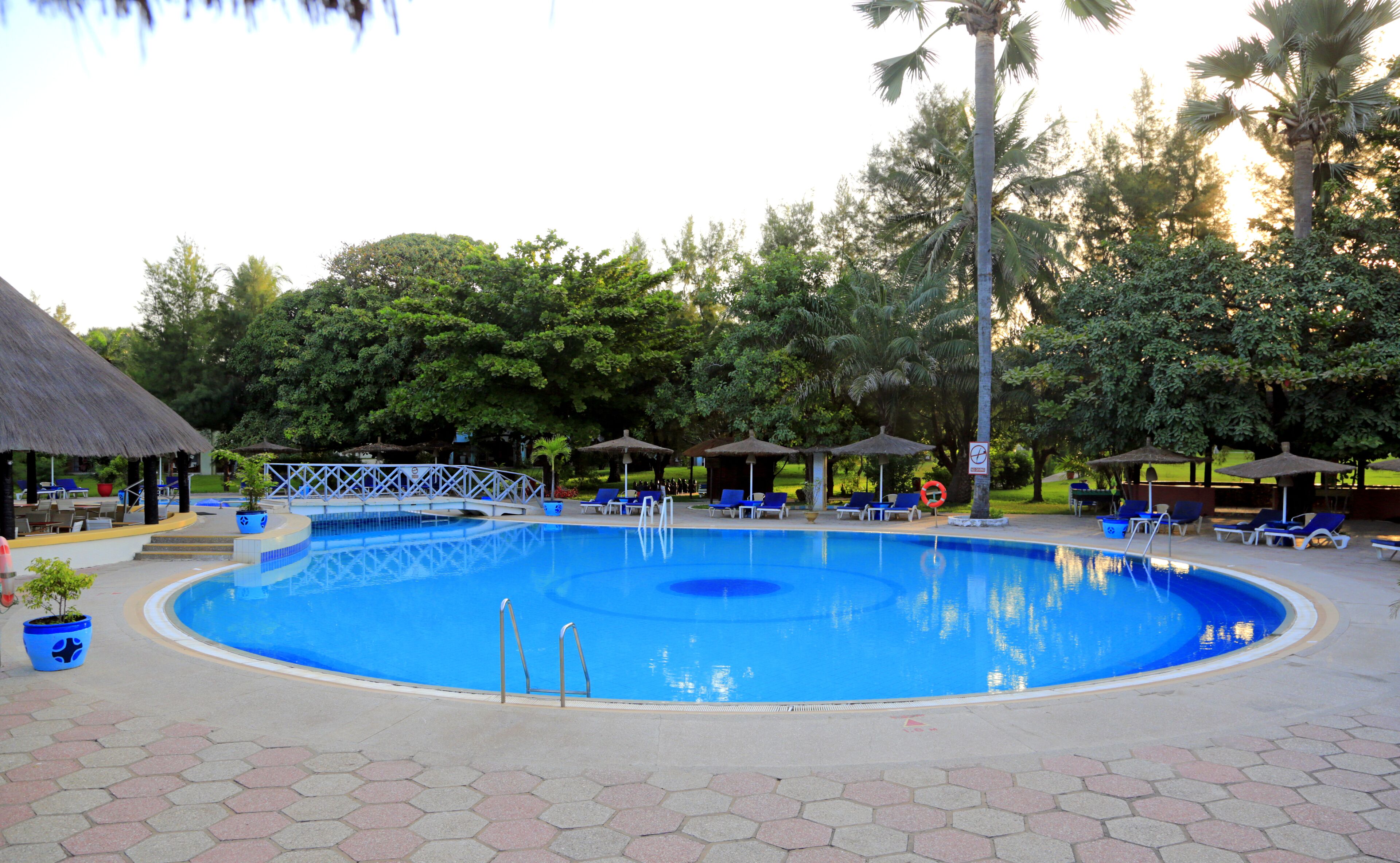 Una piscina con sillas de descanso y sombrillas, rodeada de árboles y una estructura con techo de paja.