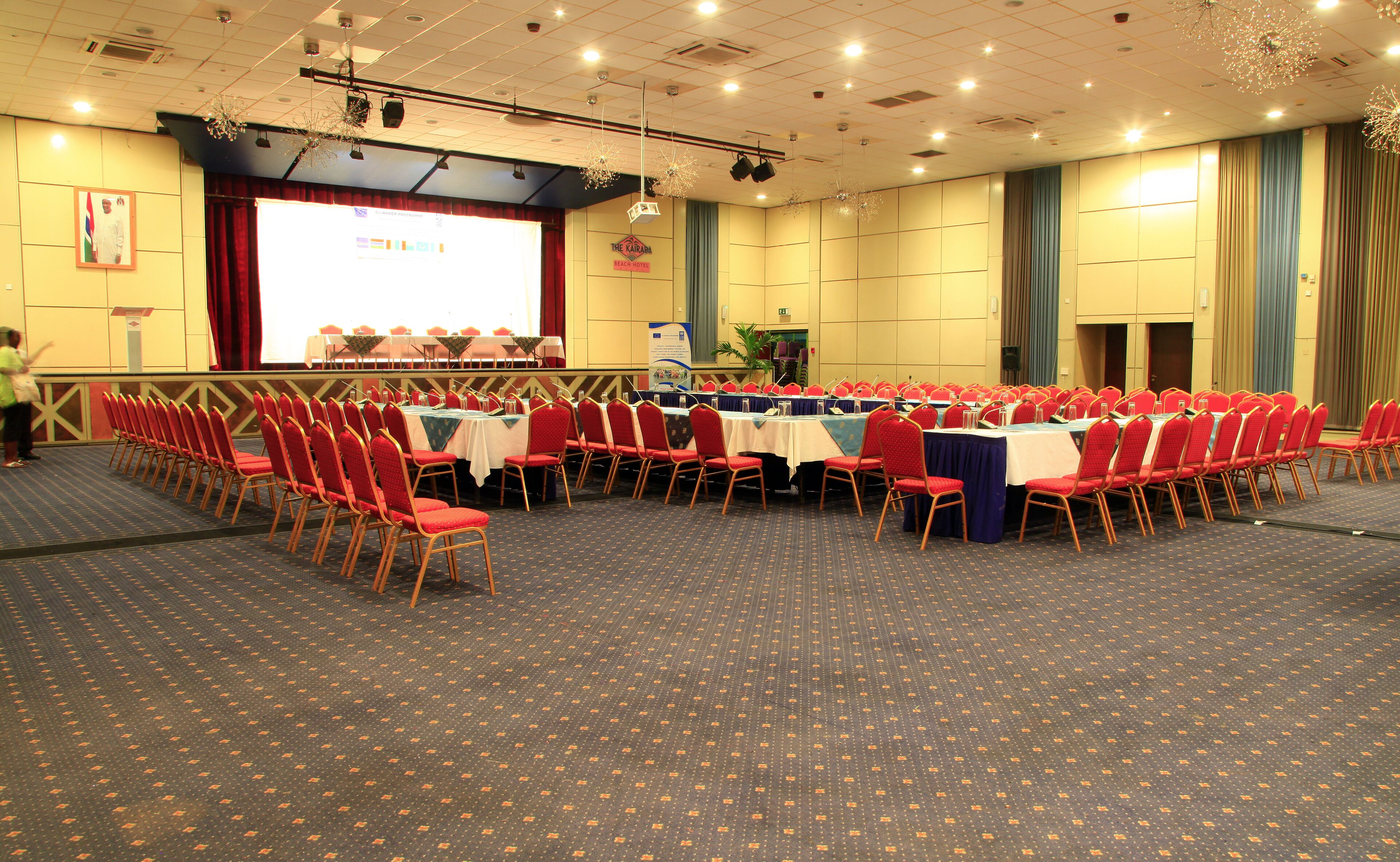 A conference room with rows of red chairs, a stage with a screen, and a carpeted floor.