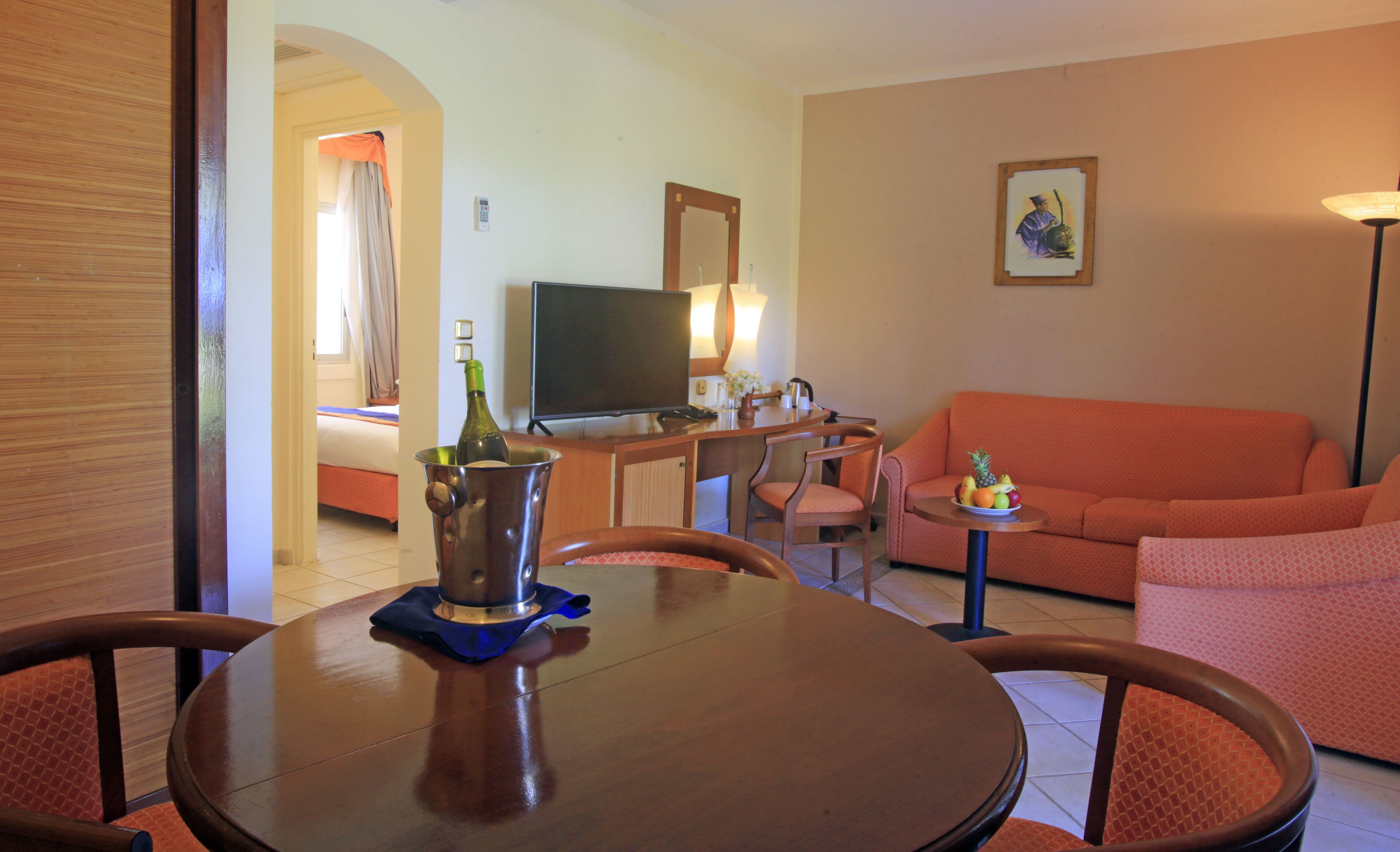A hotel room with a wooden dining table, a television, a sofa, and a small table with a fruit bowl.