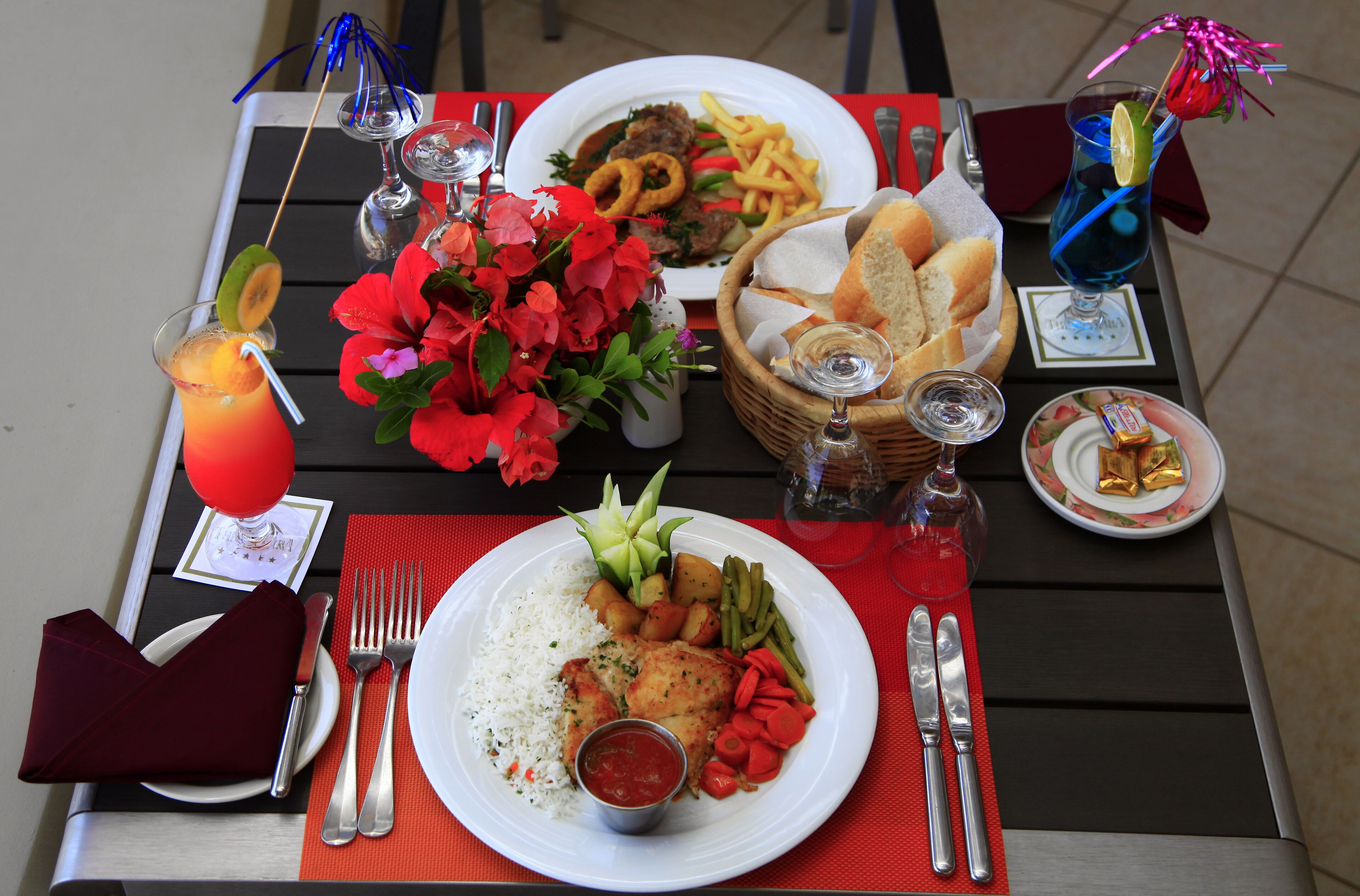 A table set with a meal including rice, vegetables, and a chicken dish, accompanied by drinks and a basket of bread.