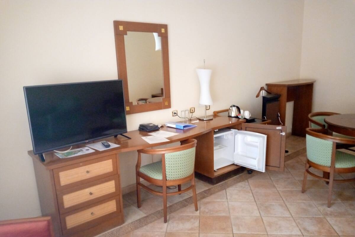 A compact hotel room with a flat-screen TV, a small refrigerator, a desk with a lamp, and a dining area with chairs.
