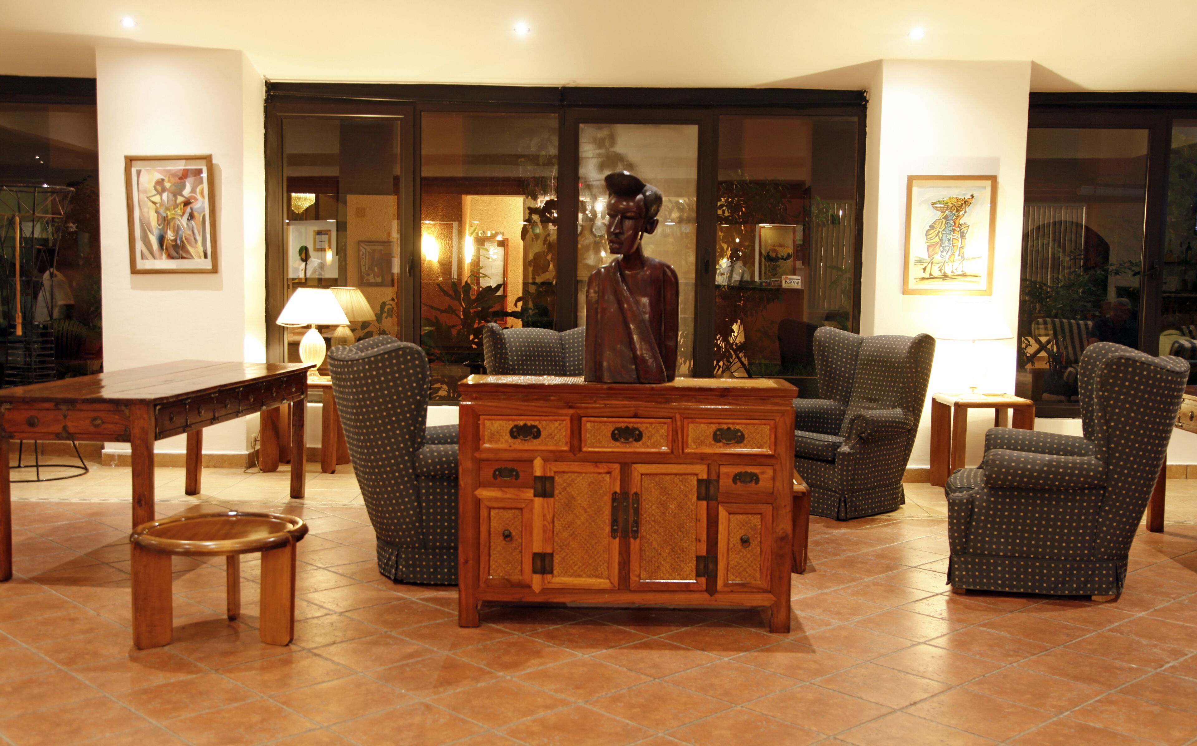 Un salon bien éclairé, avec une table basse en bois, deux fauteuils, un canapé et une table à manger. Les murs sont ornés d’œuvres d’art encadrées, et une grande fenêtre reflète la lumière.