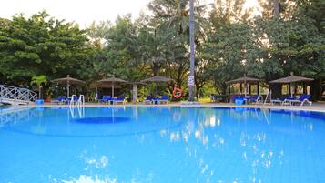 Una piscina con baldosas azules, rodeada de sillas de descanso y sombrillas, con árboles al fondo.