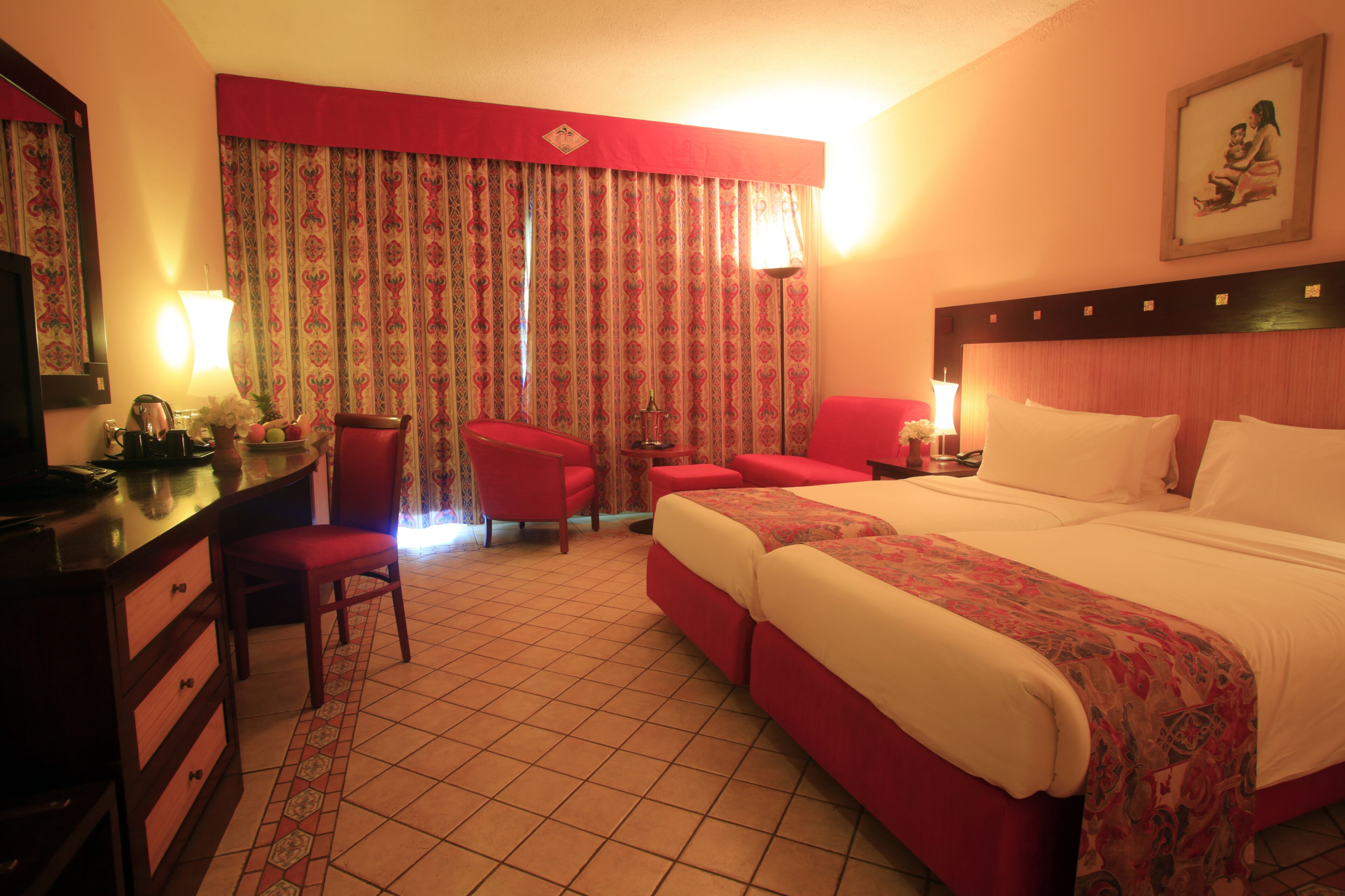 A hotel room with a large bed, two red chairs, a desk with a lamp, and a window with patterned curtains.