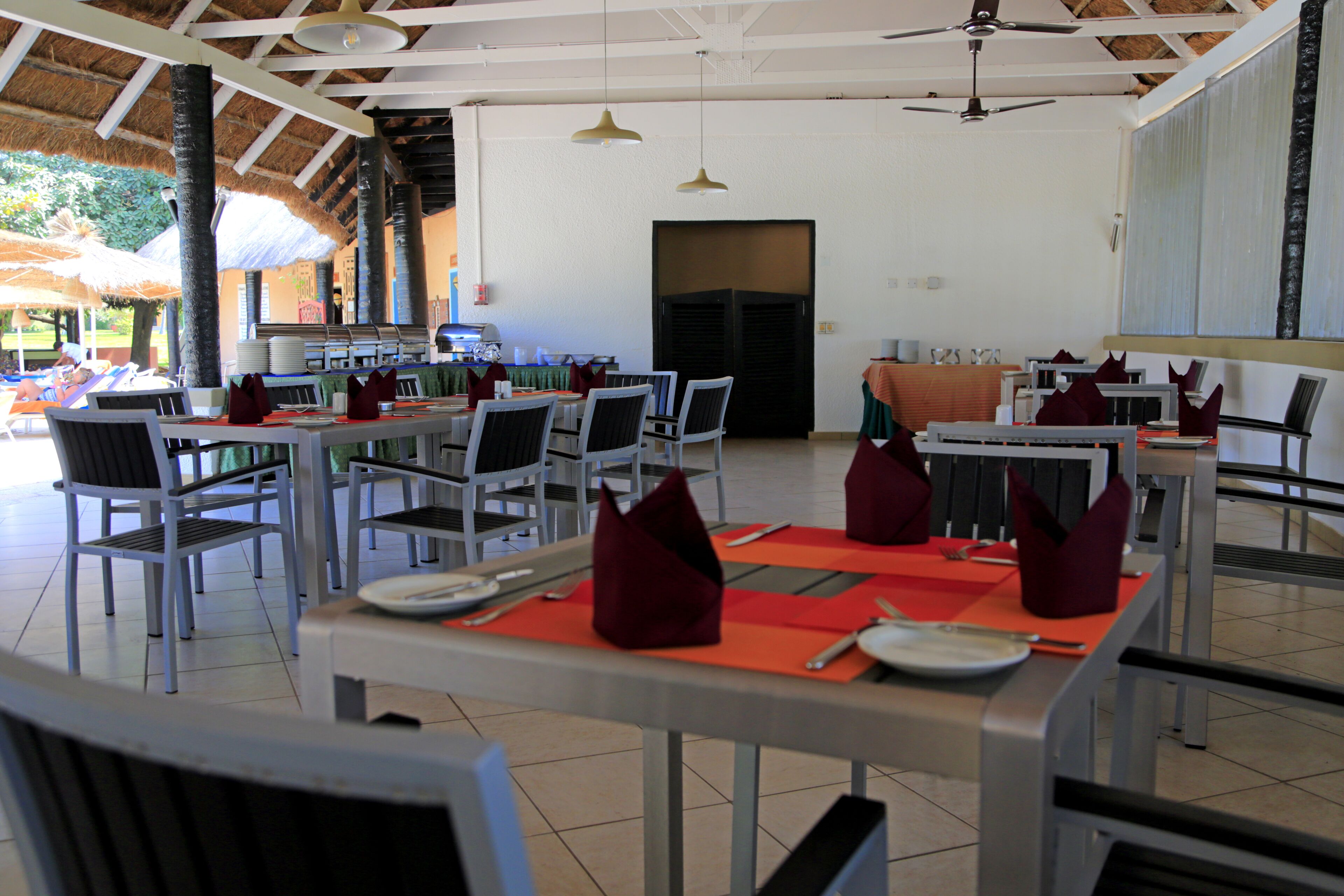 A restaurant with multiple tables and chairs, a ceiling fan, and a thatched roof.