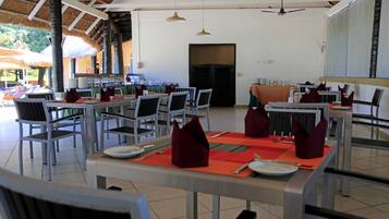 A restaurant with multiple tables and chairs, a ceiling fan, and a thatched roof.
