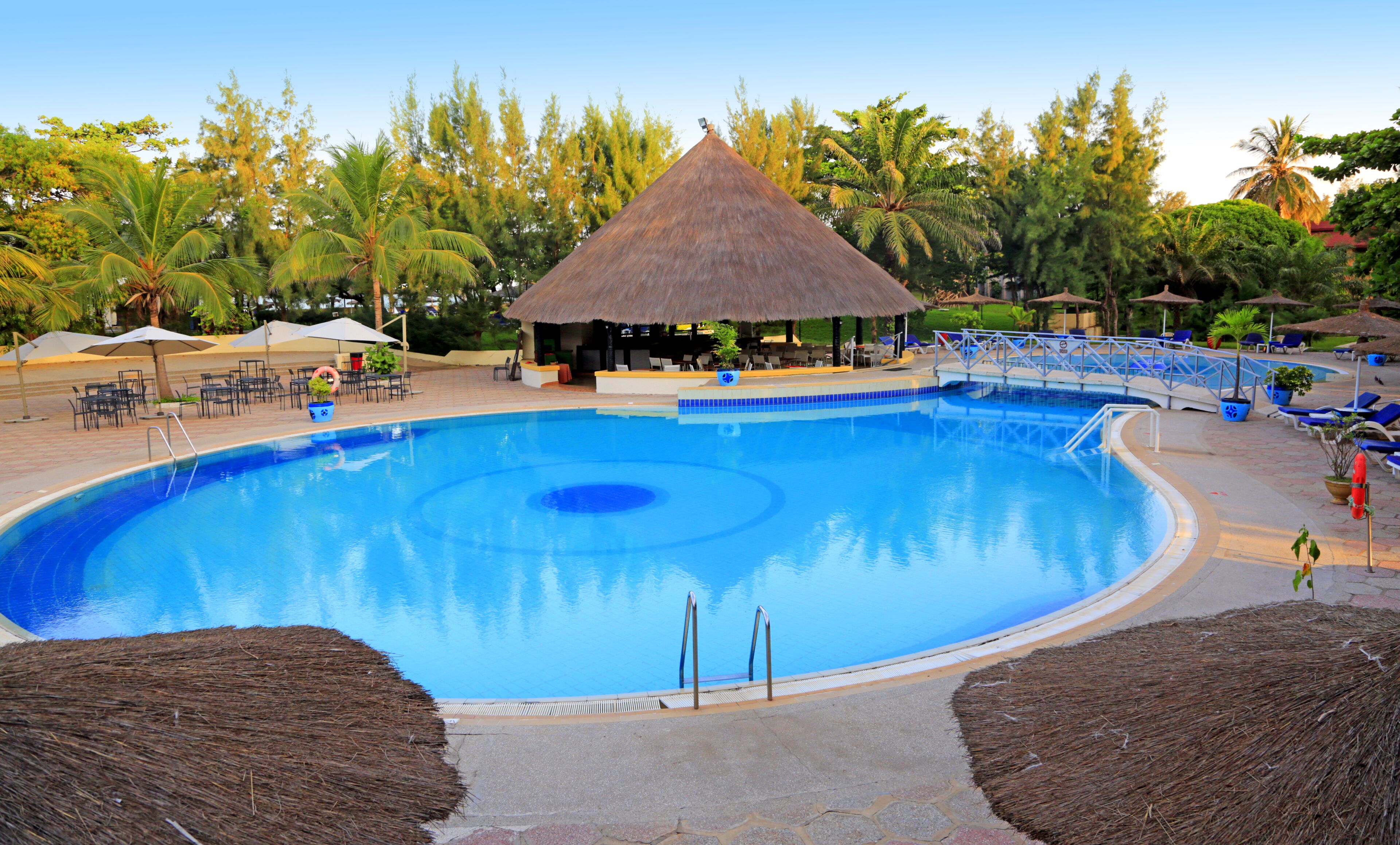 Une grande piscine avec une structure centrale dotée d’un toit de chaume, entourée de chaises longues et de parasols.
